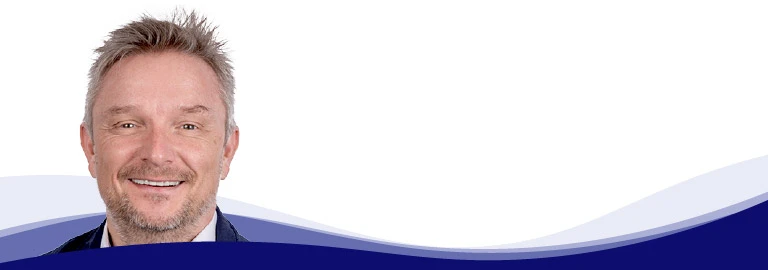 Holger Geck, Leiter Marketing und Produktdatenmanagement bei Gust. Alberts, lächelt in die Kamera. Er hat kurzes, graublondes Haar und einen kurzen Bart. Er trägt ein weißes Hemd und ein dunkles Sakko. Im Hintergrund ist eine geschwungene Wellenform in Weiß, Hellblau und Dunkelblau zu sehen.