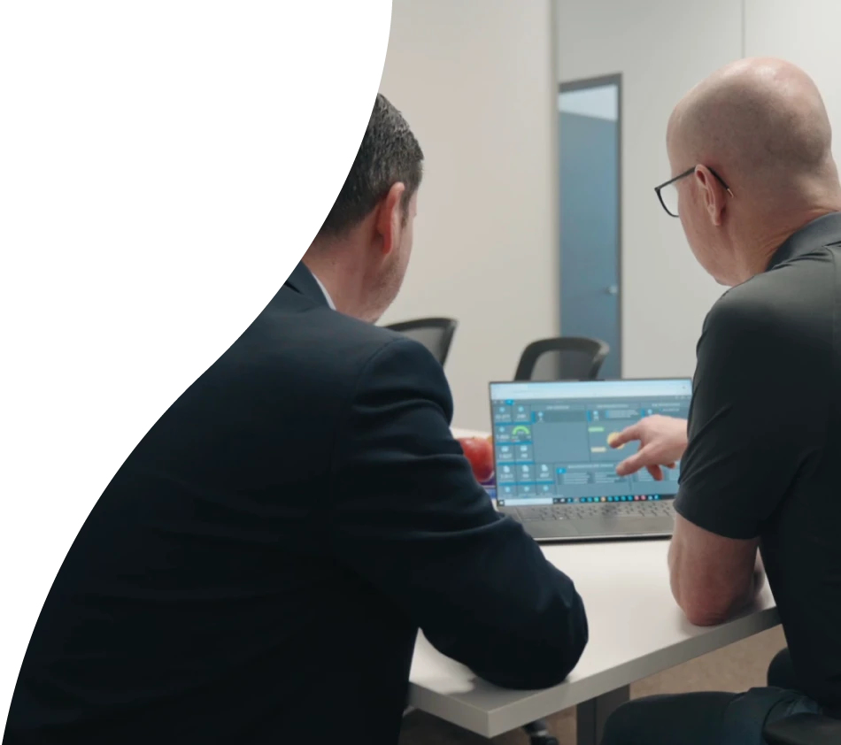 Zwei Männer sitzen mit dem Rücken zur Kamera in einem Besprechungsraum eines Büros. Auf dem Tisch vor Ihnen steht ein Laptop mit dem geöffneten Dashboard von crossbase. Der Mann rechts zeigt auf den Display und trägt eine Brille.