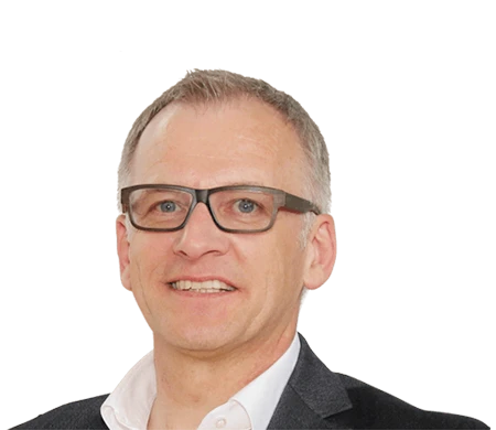 Ein Mann mittleren Alters blickt lächelnd in die Kamera. Er hat kurzes, graues Haar und trägt eine dunkle Brille mit markantem, rechteckigem Rahmen. Er ist in ein weißes Hemd mit offenem Kragen und ein dunkles Sakko gekleidet.