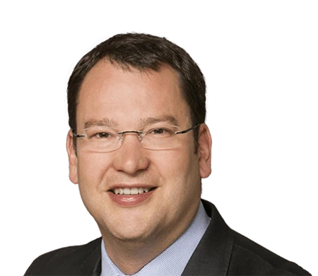 Dirk Amann, CTO Managing Director at i.safe MOBILE, looks into the camera with a smile. He is wearing rimless glasses, a light blue shirt and a dark jacket. He has short, dark brown hair and a natural complexion.