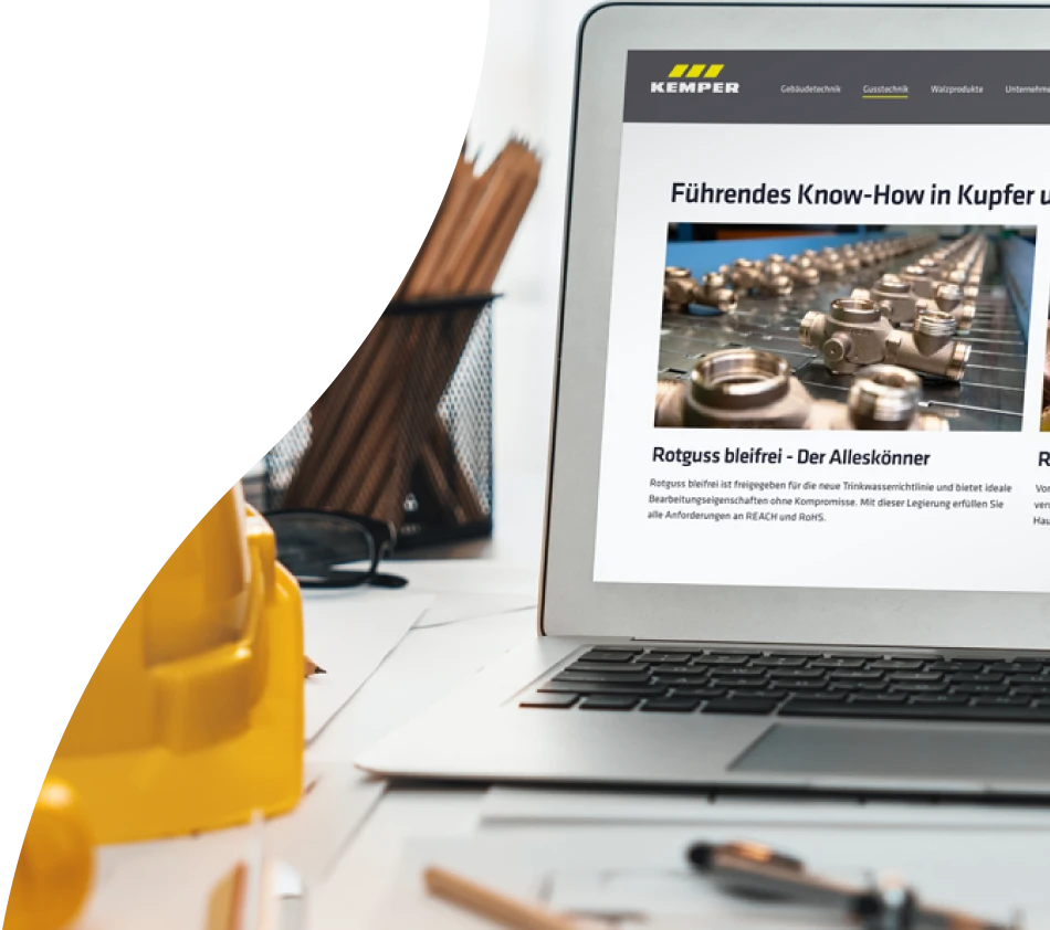 An open laptop on a table shows the screen of the KEMPER online store, which was implemented with crossbase. On the table are rolled-up plans, a hard hat for construction sites, a plant, a pair of glasses and a pencil cup.