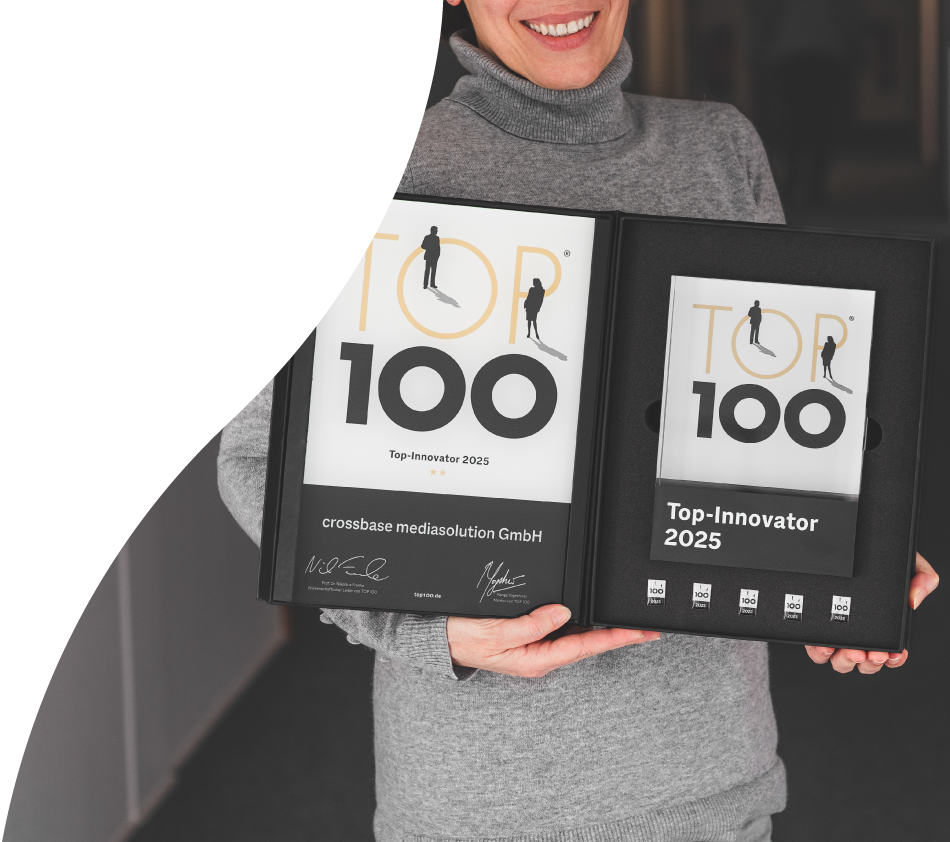A woman with a gray top holds the signed TOP 100 award for crossbase in her hands and shows it to the camera. Her head is not quite visible but she is smiling broadly.