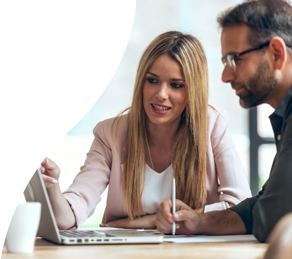 Deux personnes, une femme et un homme, sont assises ensemble à une table et regardent avec concentration l'écran d'un ordinateur portable. La femme montre l'écran et explique quelque chose pendant que l'homme prend des notes. Les deux semblent plongés dans leur travail ou dans une présentation.