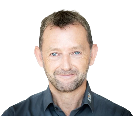 Achim Angel, directeur de KiM GmbH - Member of 4PACE, sourit aimablement à la caméra. Il a les cheveux bruns courts, une courte barbe et porte une chemise gris foncé avec un col légèrement ouvert. En arrière-plan, une forme ondulée incurvée en blanc, bleu clair et bleu foncé confère à la photo un design moderne.
