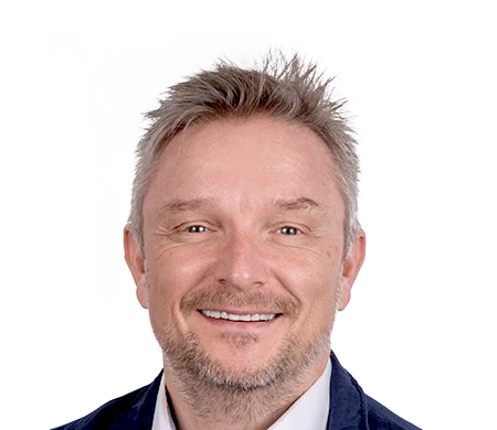 Holger Geck, directeur du marketing et de la gestion des données produits chez Gust. Alberts, sourit à la caméra. Il a les cheveux courts, blond grisâtre et une courte barbe. Il porte une chemise blanche et une veste sombre. En arrière-plan, on peut voir une forme de vague incurvée en blanc, bleu clair et bleu foncé.