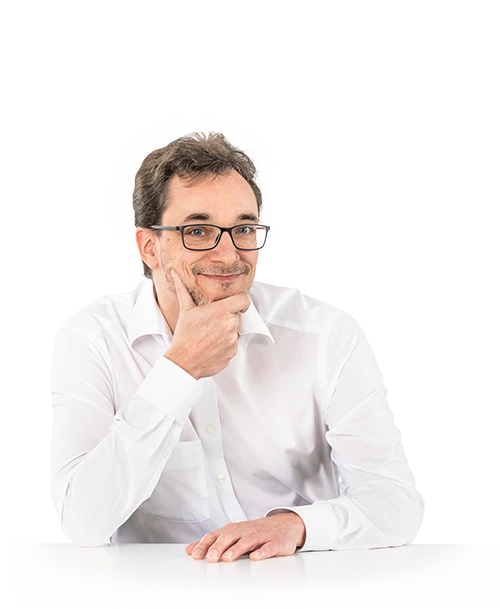 Jean-René Thies se tient à une table, le menton appuyé sur une main. Il porte des lunettes et une chemise blanche et regarde la caméra en souriant.